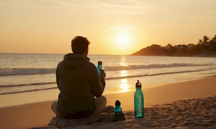 Persona sentada junto a la playa con botellas de agua a mano, acompañada de imágenes de naturaleza, puestas de sol y refrescante destinos de verano.