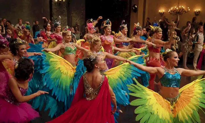 Un grupo de intérpretes en trajes de colores y maquillaje, con alas de colores brillantes en sus espaldas, realizando una rutina de baile celebratorio ante un público.