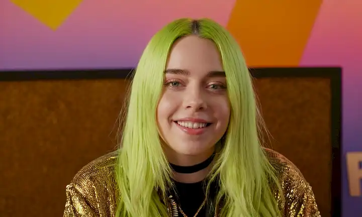 Billie Eilish sonriendo delante de su placa de nombre con fondo colorido y gestos tic.