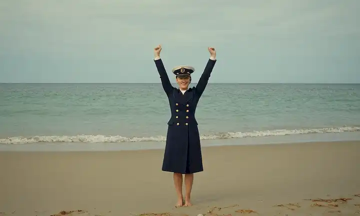 Persona usando un sombrero naval y de pie en una playa mientras rodeado de agua con sus brazos levantados en la victoria. El nombre está escrito debajo en letra script.