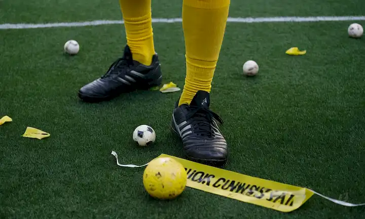 Campo de fútbol césped artificial, zapatos desgastados, cinta de precaución, piezas de goma crumbly, guantes médicos, y un jugador de aspecto preocupado en el centro, rodeado de bolas dispersas y equipo roto, con un toque de desuso y abandono en el fondo