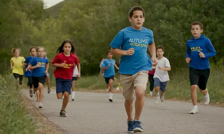 Los niños que corren junto a los pares mientras un grupo de maestros y consejeros observan, lo que simboliza el apoyo para superar los desafíos que enfrentan las personas con autismo. El texto está en español con traducción al inglés: Ejercicio: Una herramienta esencial para desarrollar el autismo: desafío y oportunidad en la Escuela Leo Kanner.
