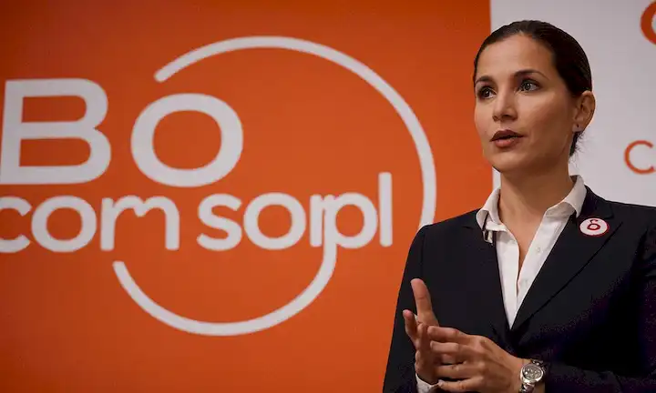Persona de pie frente a un logotipo de B Corporation mientras explica que son parte de un movimiento que cambia el sistema económico. (En inglés, esto podría ser: Una persona comparte su participación con B Corporations - una organización que busca transformar el entorno empresarial.)