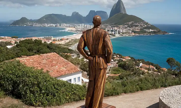 Escultura de gran escala hecha de madera reciclada por el artista brasileño Henrique Oliveira de pie frente a edificios con el continente europeo y el océano en el fondo.