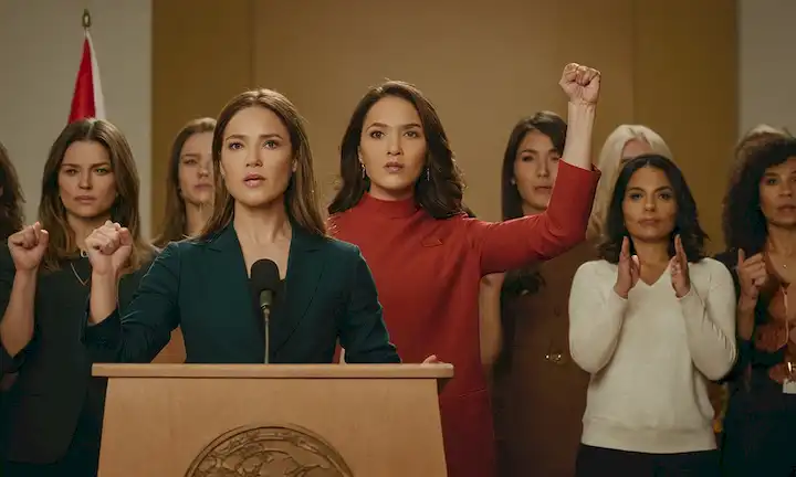Mujer detrás de un podio con banderas, acompañada por otras mujeres levantando sus puños en solidaridad. Un caption reads Unifying the Fight for Womens Rights at Global Level.