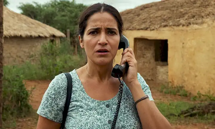 Una madre española audaz y empoderada que habla en voz alta con determinación por teléfono, transmitiendo urgencia y preocupación por sus hijos en África oriental. El fondo podría representar un pueblo bullicioso o sala educativa de su historia. Las palabras Un llamado a la acción: No te quedes de brazos cruzados traducen a una llamada a la acción: No te quedes de lado - una llamada a la acción.