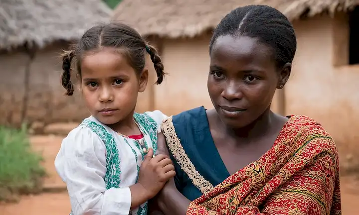 Una joven en un pueblo local con la mano de su madre, ambos con ropa tradicional. Una mujer llamada María Galán, una mujer española que viajó a Uganda hace unas décadas y desde entonces ha hecho una vida allí alimentando y criando niños. La frase sugiere su historia de triunfo contra todas las probabilidades, probablemente arraigadas en sacrificio personal, y adaptación cultural a la realidad africana.