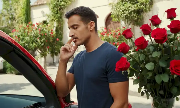 Hombre con dedo en nariz y rosas rojas en fondo de arreglos florales, de pie junto a un coche. En español.