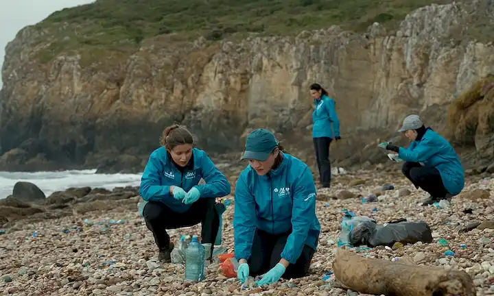 Equipo de investigadores que llevan equipo de seguridad trabajando en la orilla de las zonas costeras, discutiendo desechos de botellas de plástico descartadas con caras de fondo. El subtítulo inglés dice: El papel de la investigación de la pellets de botella de plástico: Playas de Galicia y Asturias todavía afectados.