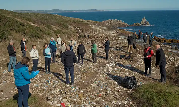 Un grupo de participantes interesados se reunieron por un tramo de costas dañadas cerca de las costas de Galicia y Asturia, discutiendo esfuerzos de limpieza en medio de escombros dispersos en medio de pellets de plástico reciclado en el idioma inglés.
