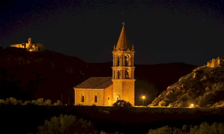 Una iglesia y su campanario se iluminaron por la noche, con canto distante acompañandola. Esto se refiere a un sitio del Patrimonio Mundial de la UNESCO, La Capilla de la Iglesia, ubicado en la ciudad de Belchite en España. La obra cuenta con sonidos misteriosos e inexplicables llamados psicofonías que han venido a representar las leyendas y misterios de las ciudades. En esta descripción, he centrado en los aspectos visuales únicos de esta estructura arquitectónica en medio de un telón de sonido y luz que encapsula su historia y significado.