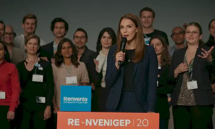 Mujer que habla en el escenario mientras está rodeada de colegas y participantes frente a una imagen o logo que muestra el nombre Reinventa-Tech 2.0. La cámara también se dirige a otra mujer con un fondo técnico, ambos de pie en medio de una reunión de entusiastas tecnológicos animando con emoción en sus ojos.