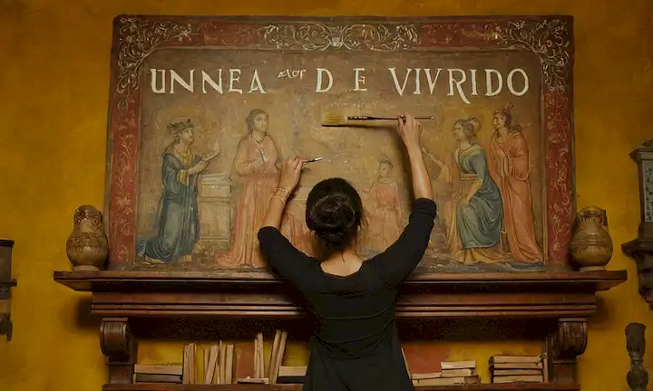 Mujer de pie sobre una antigua estantería, sosteniendo un pincel mientras el título Una Enciclopedia para Desafiar El Olvido aparece en negrita y femenina escribiendo detrás de ella. El fondo cuenta con una multitud diversa de artistas femeninos, cada uno trabajando hábilmente en su propia obra maestra. La frase La Historia del Arte con rostro de mujer: Un Viaje A Thru Es De 500 Muchoas Artistas está acompañado por la firma de artistas junto a su pintura en caligrafía manuscrita.