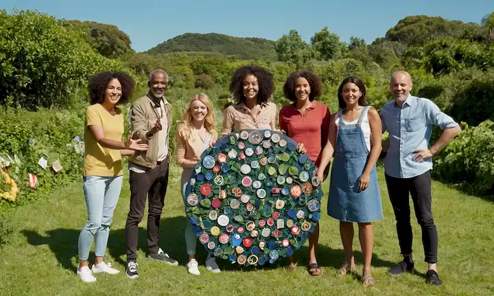 3-5 personas, diversos grupos de edad y habilidades, sonriendo, de pie frente a exuberante vegetación, rodeado de símbolos de reciclaje y materiales reciclados, agarrando manos o formando un círculo, con un cielo azul y paneles solares en el fondo