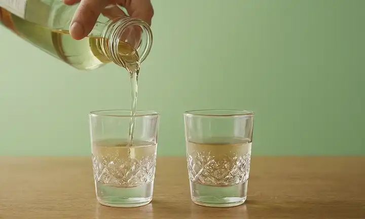 Persona vierte vinagre blanco en un vaso con otro vierte añadiendo agua, luego mezclando los dos. Texto en una fuente pequeña. Una alternativa ecológica al vinagre blanco para refrescos. En inglés según lo soliciten los usuarios.