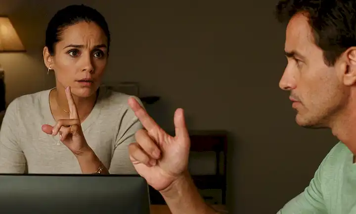 El hombre mirando fijamente a su pantalla del ordenador mientras la mujer da consejos con gestos de mano. Texto español superpuesto. ¿Es saludable o una adicción? ¡Averiguar descubriendo!.