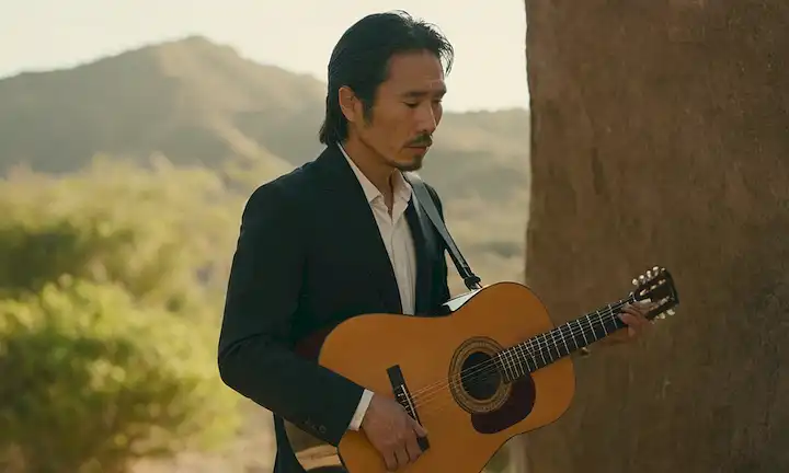 Hombre japonés de pie fuera con una guitarra, mirando pensante mientras explica el proceso artístico detrás de su pasión por el flamenco.
