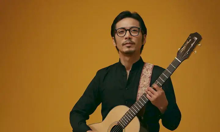 Persona de pie delante de un telón de fondo con traje de guitarra y danza, usando gafas con el nombre Yoichiro Yamada. El subtítulo se refiere a su viaje para practicar flamenco a pesar de los obstáculos. En español, potencialmente Yoichiro Yamada: Un ejemplo de superación de Yoichiro Yamada: La obsesión por el flamenco que lo llevó a la calle.