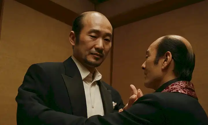 Hombre japonés de altura promedio expresando gratitud a un hombre calvo, seguido de fotos de ambos hombres juntos, y el título en una fuente audaz: Yamadas Flamenca Obsesión: El Hombre Detrás de Japón Escena de Stripping.